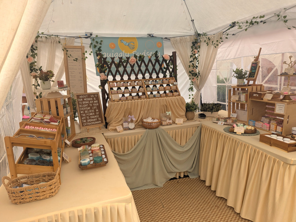 A rustic market booth set up inside a white tent, featuring draped beige tablecloths, wooden shelving, baskets, and various artisanal goods and signs.