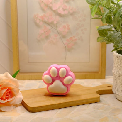 A pink and white paw-shaped soap, displayed on a wooden cutting board with a peach rose, a framed print, and a potted plant in the background.