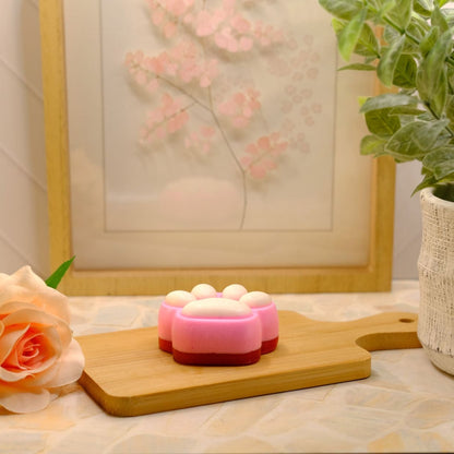 A pink and red paw-shaped soap with white bubble details sits on a wooden cutting board, styled with a peach rose, a potted plant, and a framed print in the background.