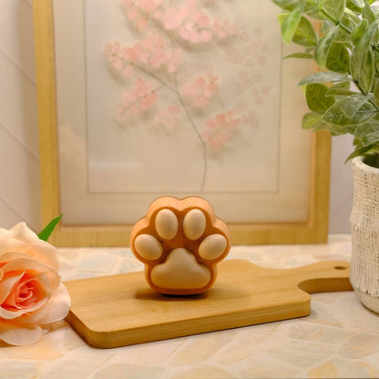 A small orange, brown and white paw-shaped soap sits on a wooden cutting board on a tabletop, styled with a peach rose, a potted green plant, and a framed floral print in the background.