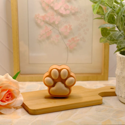 A small orange, brown and white paw-shaped soap sits on a wooden cutting board on a tabletop, styled with a peach rose, a potted green plant, and a framed floral print in the background.