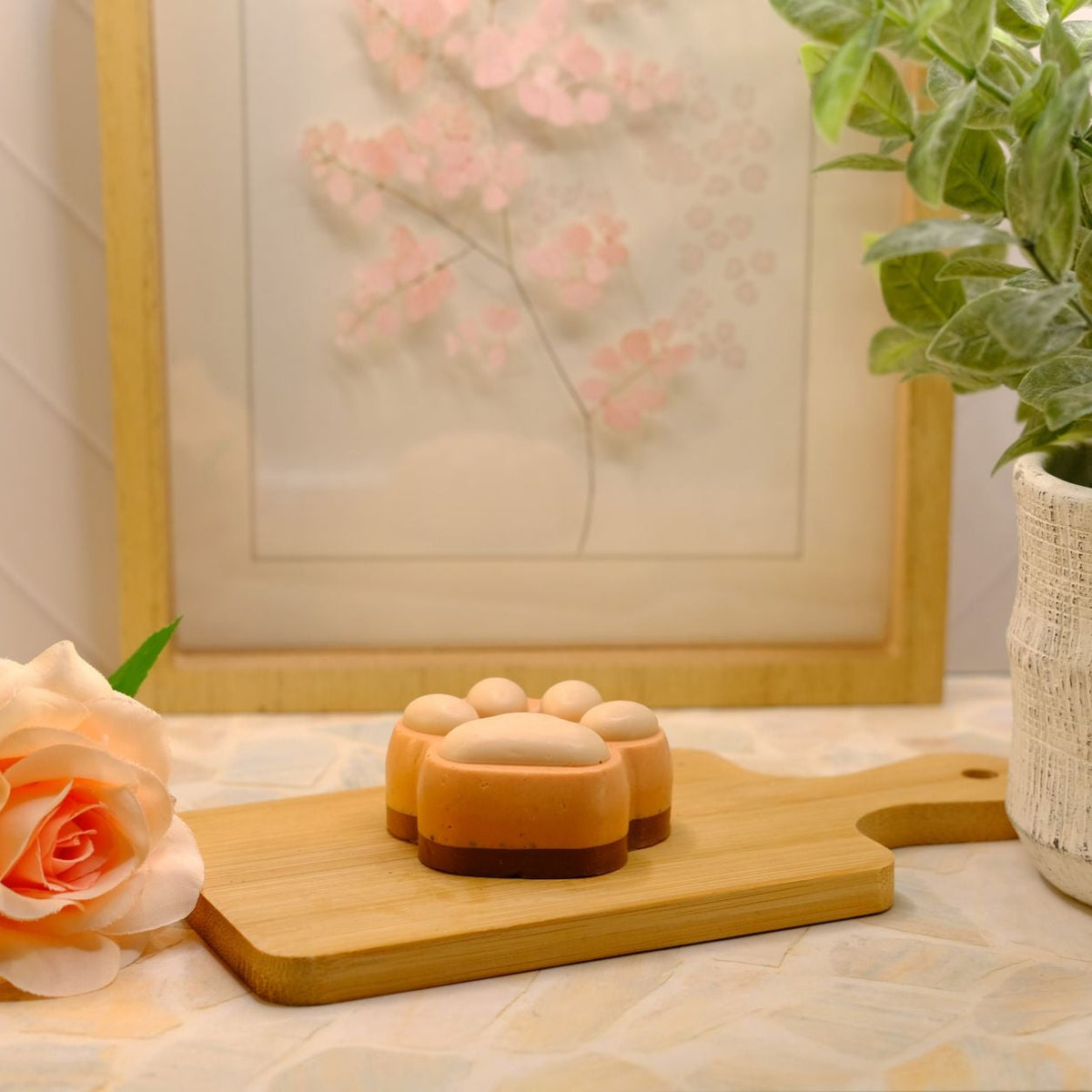 An orange paw-shaped soap bar resting on a small wooden cutting board, styled with a peach rose, a potted green plant, and a framed floral print in the background.