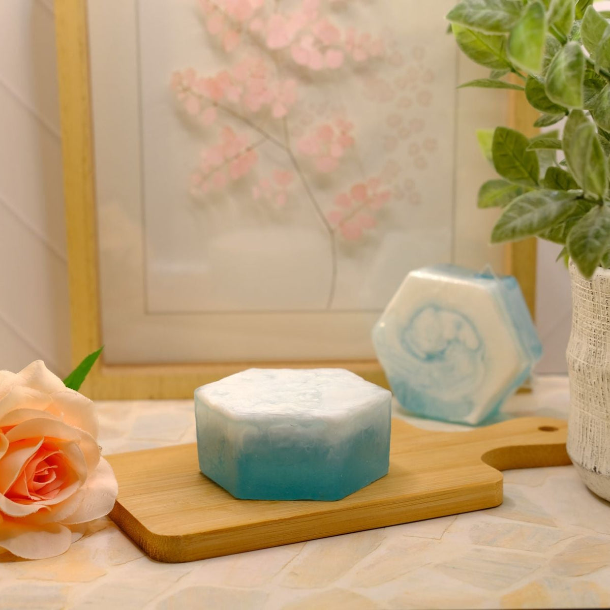 A hexagonal blue and white soap bar sits on a wooden cutting board on a tiled countertop, styled with a peach rose, a small hexagonal blue candle, and a potted green plant in the background.