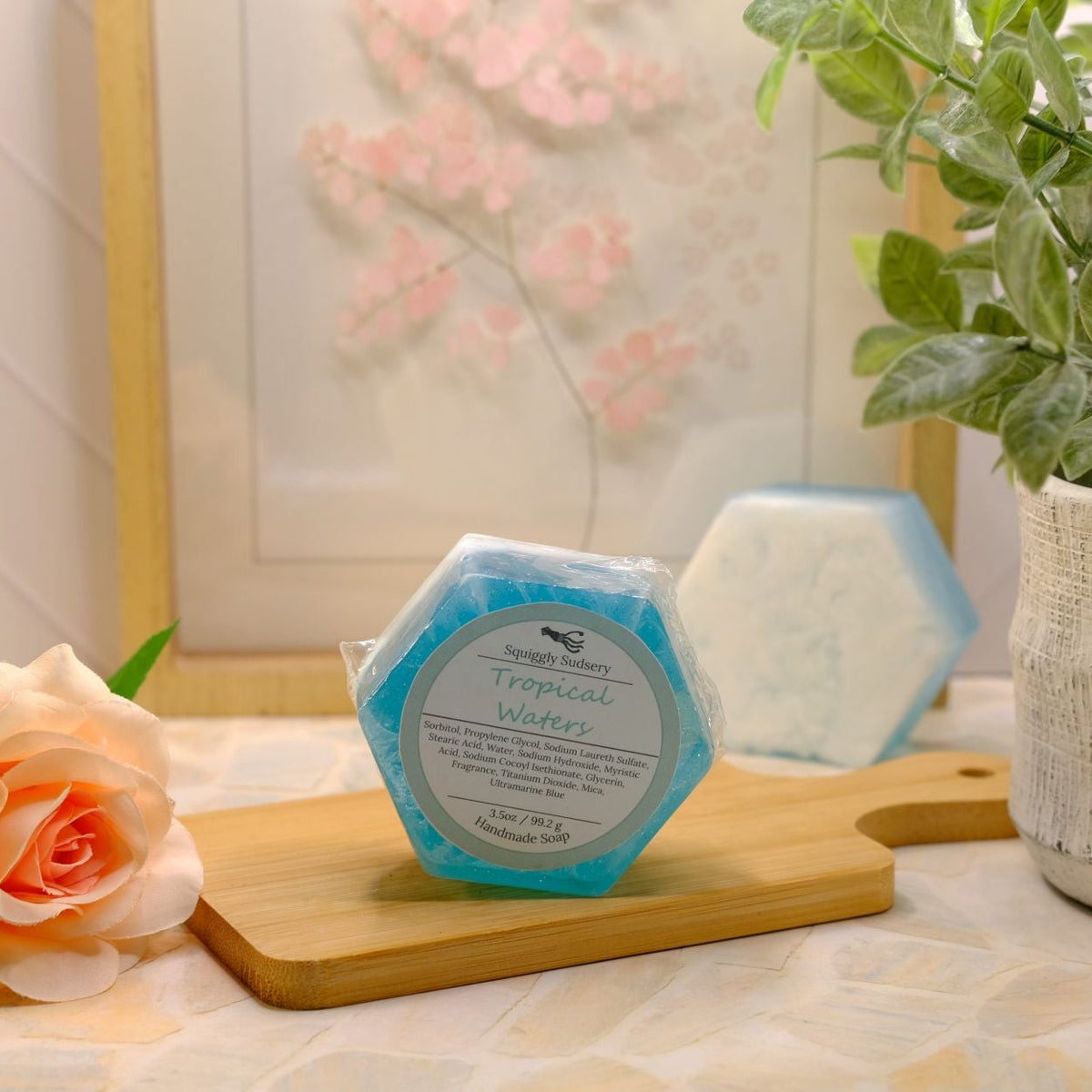 A hexagonal blue and white soap bar wrapped in clear plastic with a circular label reading 'Tropical Waters,' displayed on a wooden cutting board on a tabletop. A peach rose, a potted green plant, and a framed floral print are arranged around it in a styled indoor setting.