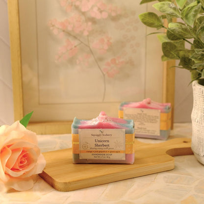 A styled product shot of a handmade soap bar labeled 'Unicorn Sherbert' by Squiggly Sudsery, resting on a wooden cutting board next to a peach rose, with a potted plant and framed art in the background.