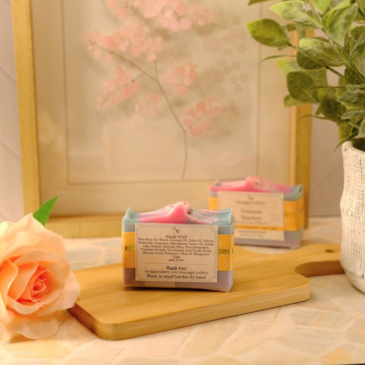 Two handmade soap bars wrapped in kraft paper with pink ribbons, displayed on a wooden cutting board next to a peach rose and a potted green plant.