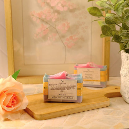 Two handmade soap bars wrapped in kraft paper with pink ribbons, displayed on a wooden cutting board next to a peach rose and a potted green plant.