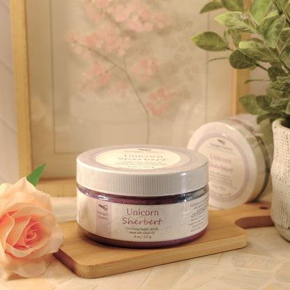 A jar of Unicorn Sherbert sugar scrub sits on a wooden block on a tabletop, styled with a peach rose, a potted plant, and a framed print in the background.