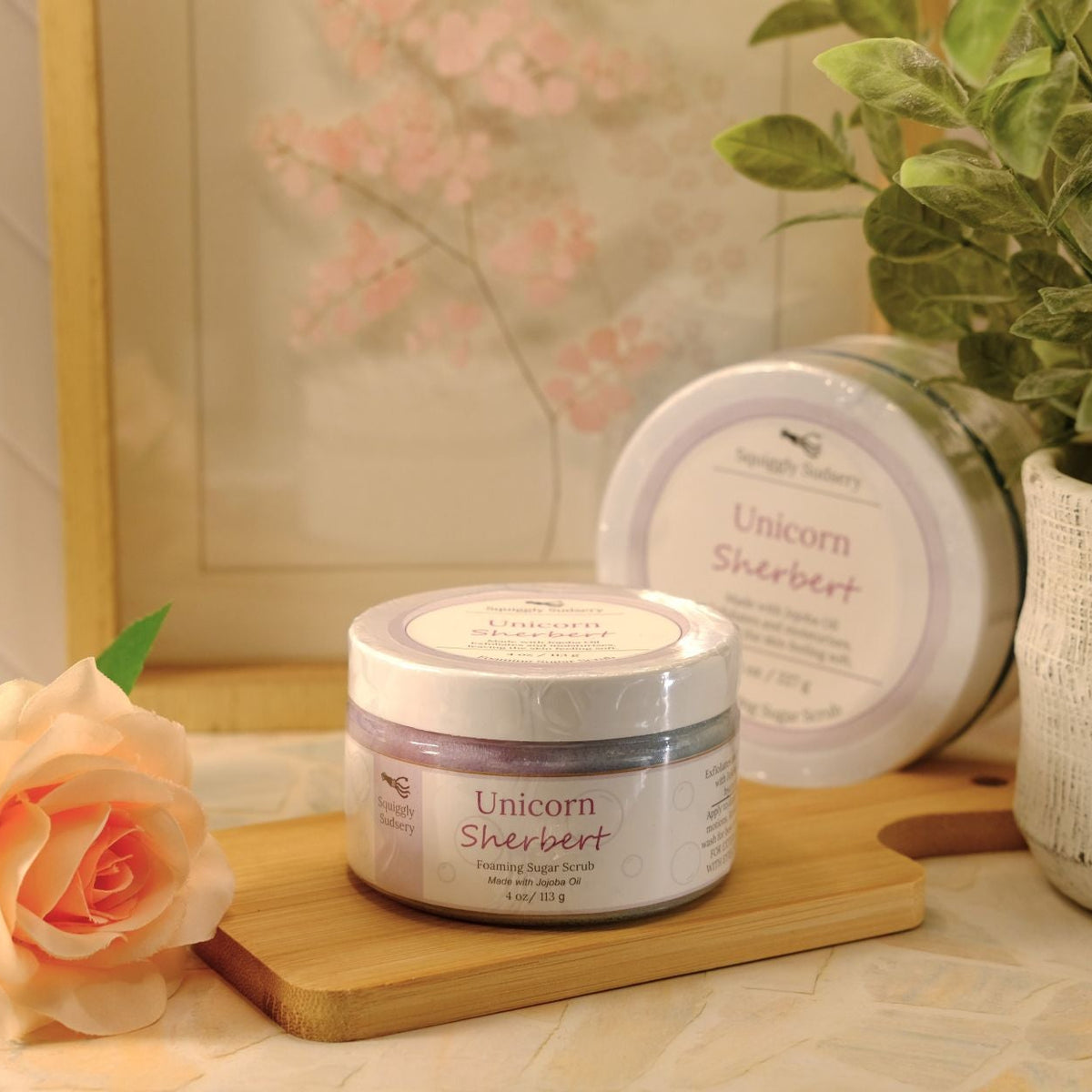 A jar of Unicorn Sherbert sugar scrub sits on a wooden block on a patterned tablecloth, styled with a peach rose, a potted plant, and a second balm tin in the background.