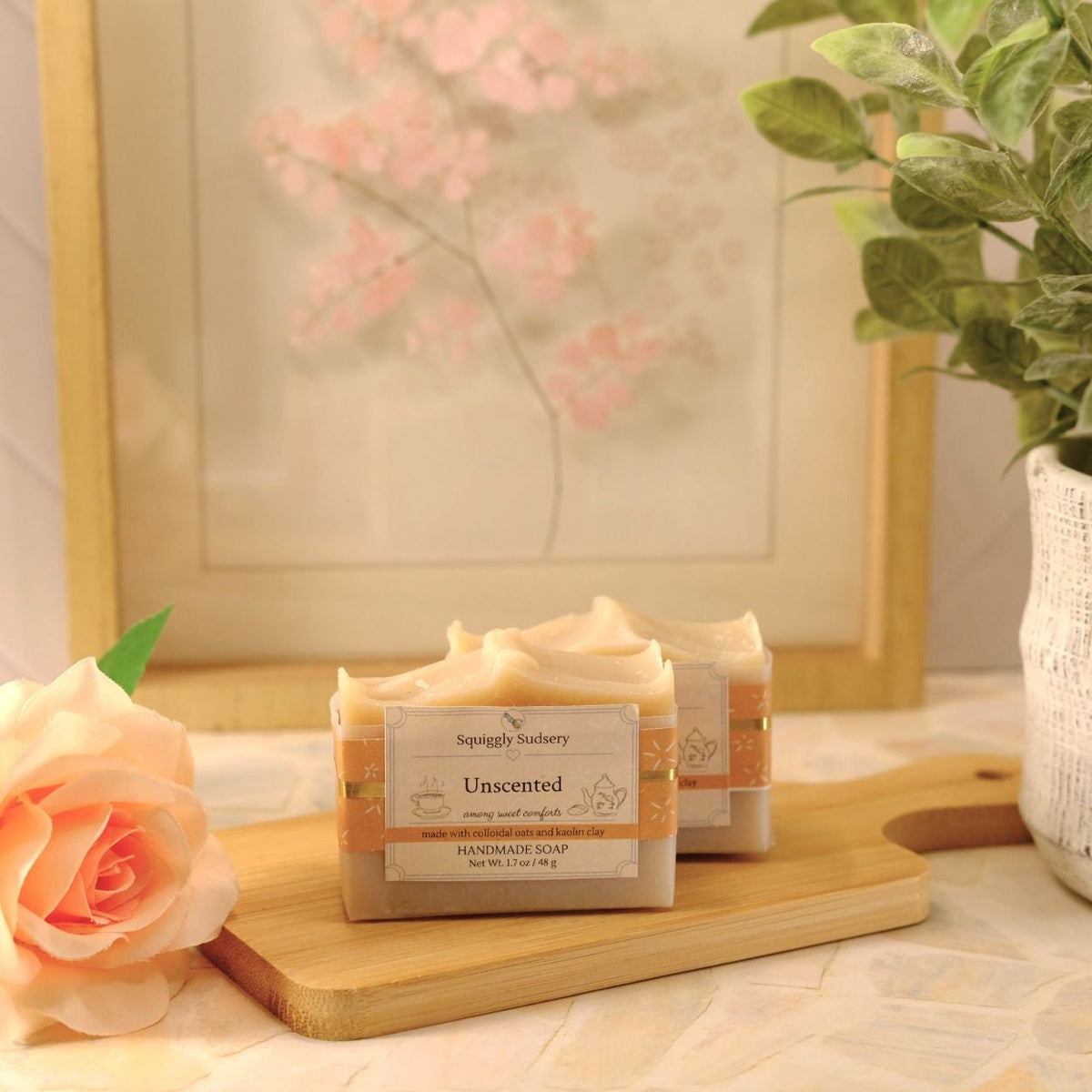 Two bars of soap labeled 'Unscented' by Squiggly Sudsery, displayed on a wooden cutting board with a peach rose and a potted plant in a softly lit setting.