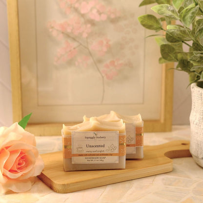 Two bars of soap labeled 'Unscented' by Squiggly Sudsery, displayed on a wooden cutting board with a peach rose and a potted plant in a softly lit setting.