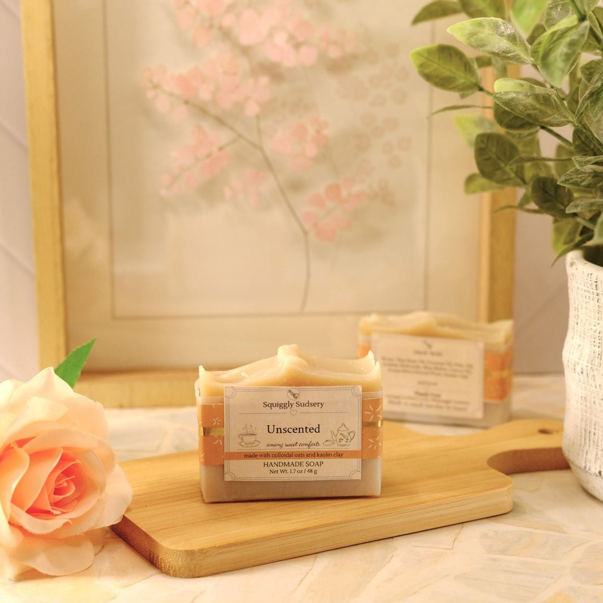A bar of soap labeled 'Unscented' by Squiggly Sudsery sits on a wooden cutting board on a tabletop, styled with a peach rose, a potted plant, and a framed print in a bright, soft setting.