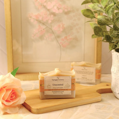 A bar of soap labeled 'Unscented' by Squiggly Sudsery sits on a wooden cutting board on a tabletop, styled with a peach rose, a potted plant, and a framed print in a bright, soft setting.