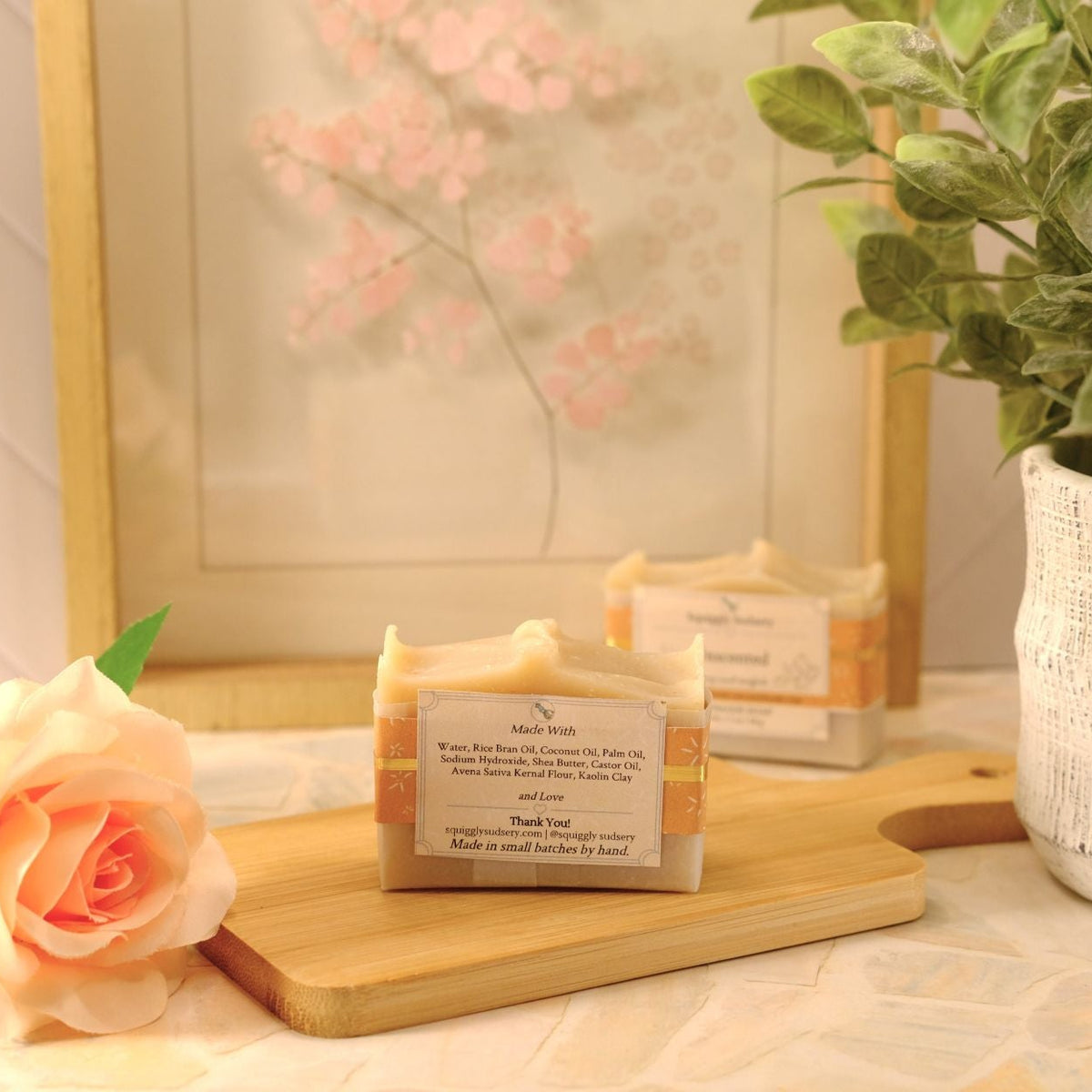 A handmade soap bar resting on a wooden cutting board, styled with a peach rose, a potted plant, and a framed print in a warm, soft-focus setting.
