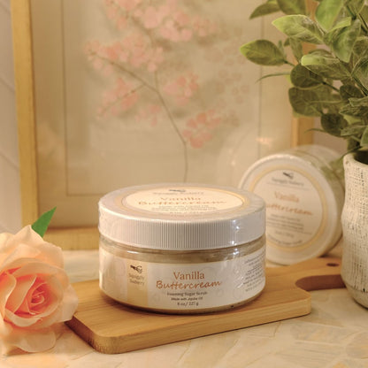 A jar of Vanilla Buttercream sugar scrub sits on a wooden cutting board on a marble countertop, styled with a peach rose, a potted green plant, and a framed floral print in the background.