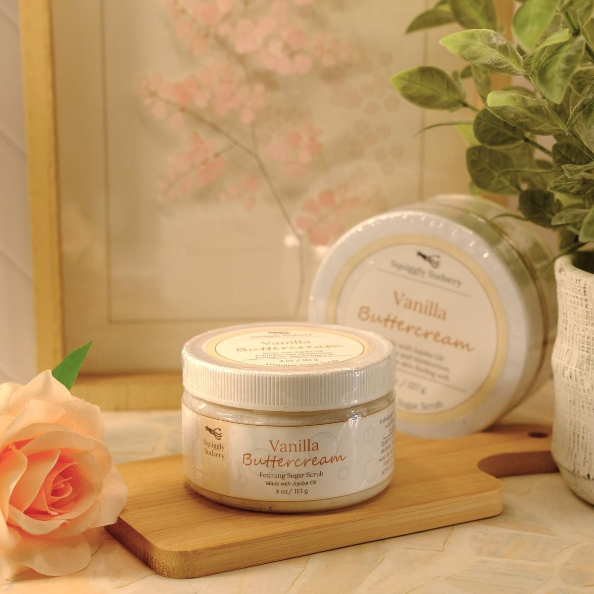 A jar of Vanilla Buttercream sugar scrub and a round jar of the same product are displayed on a wooden board, styled with a peach rose and a potted plant in a warm, soft-focus setting.