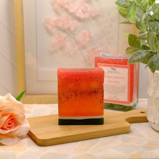 A rectangular orange-red soap with dark speckles sits on a wooden board on a patterned tabletop, with a packaged soap in the background, a potted green plant, and a peach rose.