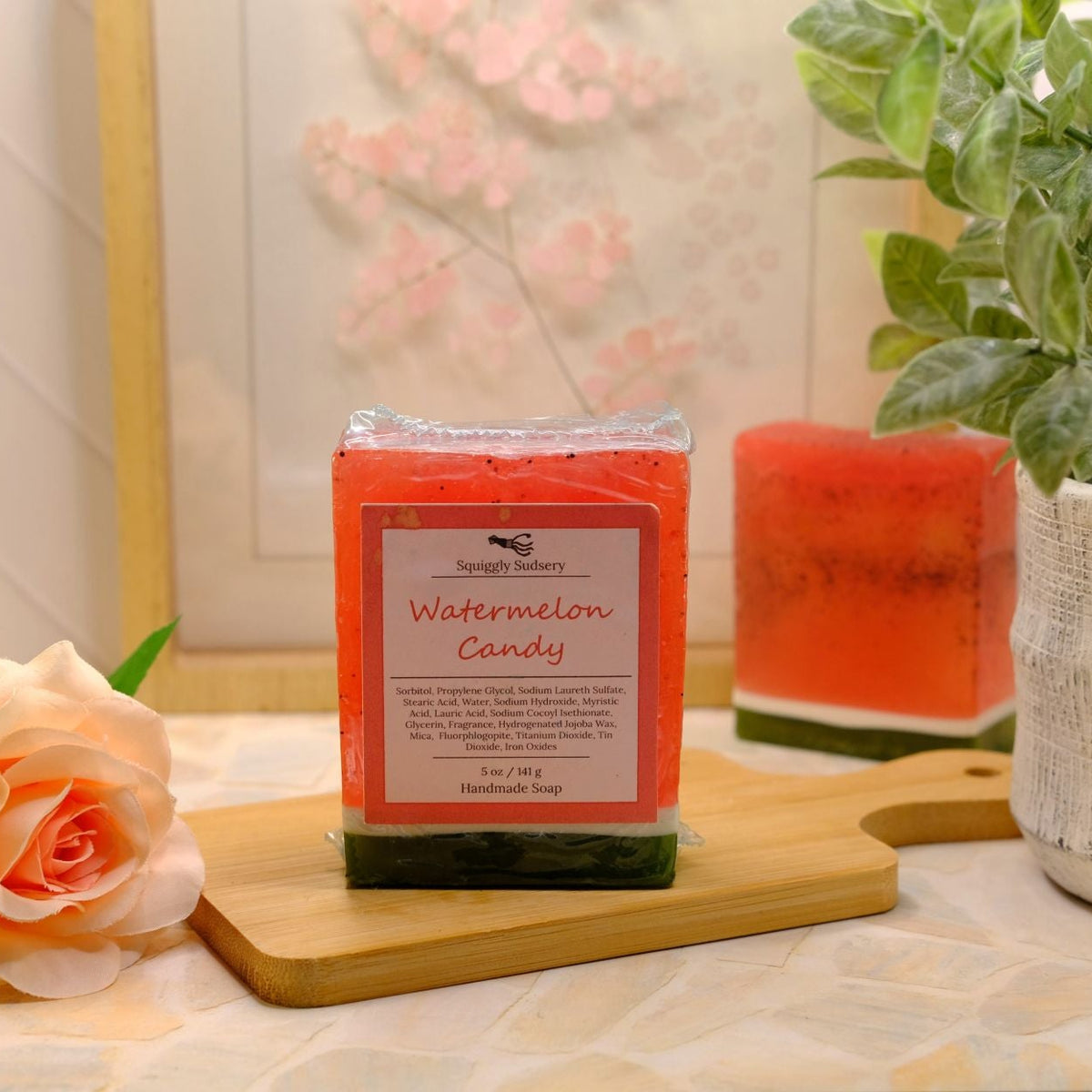 A wrapped soap labeled 'Watermelon Candy' by Squiggly Sudsery sits on a wooden board on a tabletop, styled with a rose, another candle, and a potted plant in a home-like setting.