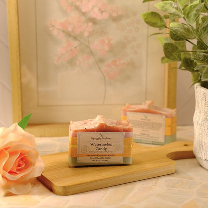 A bar of soap labeled 'Watermelon Candy' by Squiggly Sudsery, styled on a wooden cutting board next to a peach rose and a potted green plant in a warm, soft-focus setting.