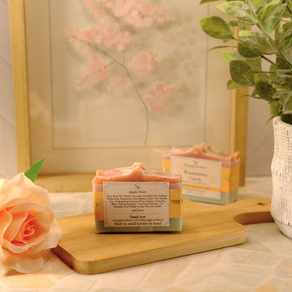 A rectangular handmade soap bar sits on a wooden cutting board, styled with a peach rose and a potted green plant in a warm, soft-focus setting.