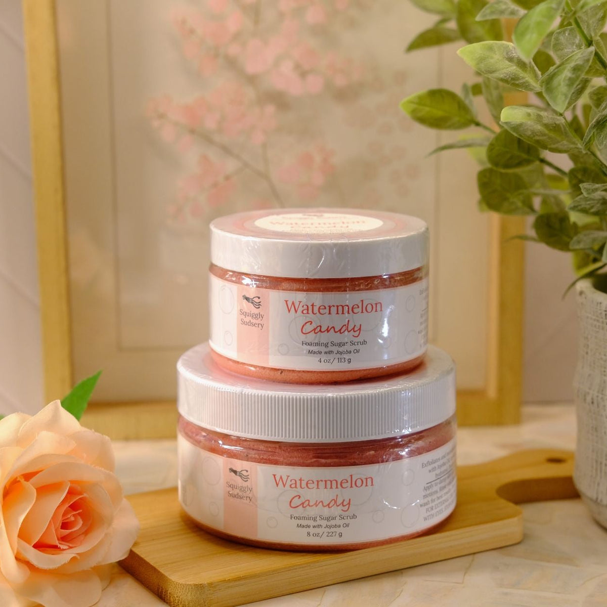 Two stacked jars of Watermelon Candy sugar scrubs by the Squiggly Sudsery on a wooden tray, styled with a peach rose and a potted green plant against a warm, neutral background.