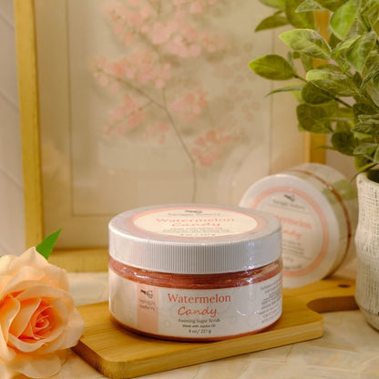 A jar of Watermelon Candy sugar scrub sits on a wooden block on a tabletop, styled with a peach rose, a potted green plant, and a framed print in the background.