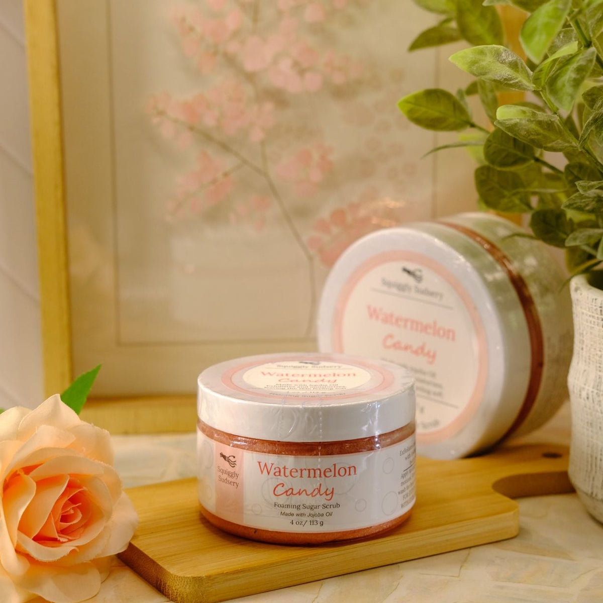 A jar of Watermelon Candy scented sugar scrub is displayed on a wooden cutting board next to a rose, with another round jar and a potted plant in the background.
