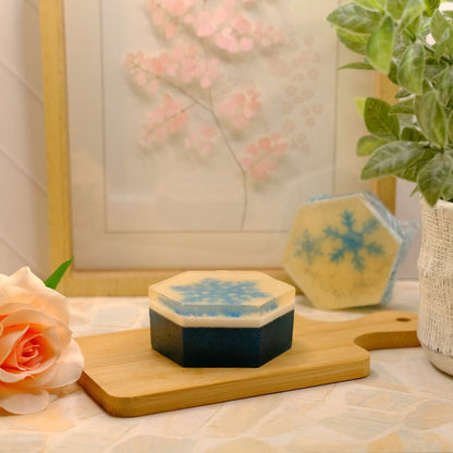 A hexagonal soap bar with a dark navy base and light blue snowflake pattern, displayed on a wooden cutting board next to a peach rose and a potted plant.