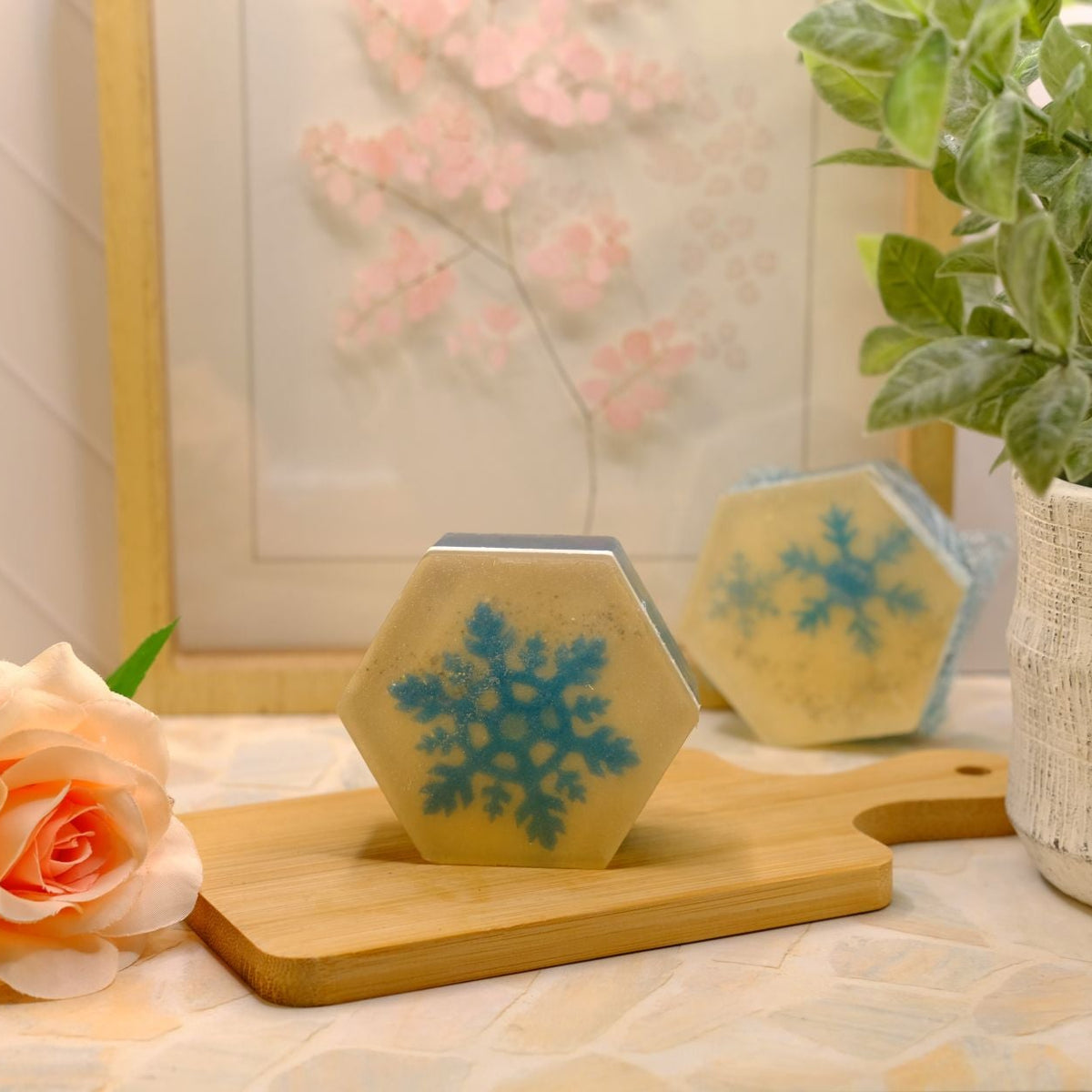 Two hexagonal soaps with blue snowflake designs resting on a wooden cutting board, styled with a peach rose and a potted green plant in a bright, warm setting.