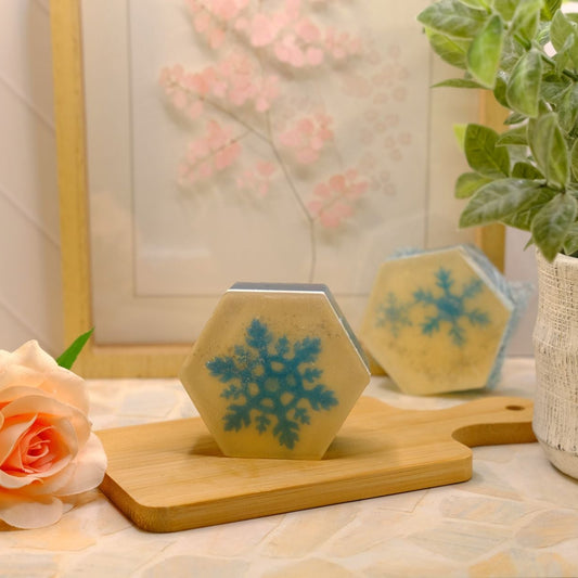 Two hexagonal soaps with blue snowflake designs resting on a wooden cutting board, styled with a peach rose and a potted green plant in a bright, warm setting.