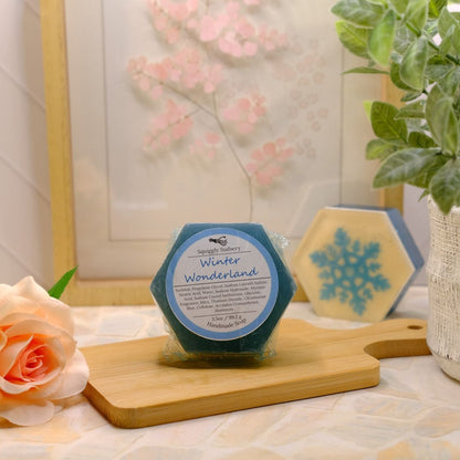 A hexagonal soap labeled 'Winter Wonderland' sits on a wooden cutting board on a tabletop, styled with a peach rose, a potted green plant, and another hexagonal item in the background.