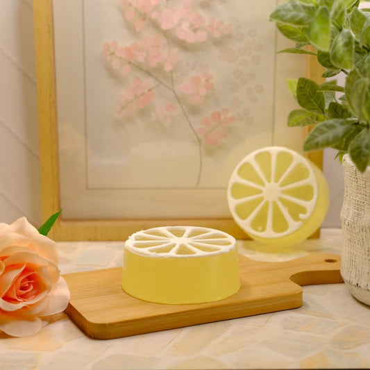 A yellow lemon slice-shaped soap on a wooden cutting board, styled with a peach rose, a potted green plant, and a framed floral print in a warm indoor setting.