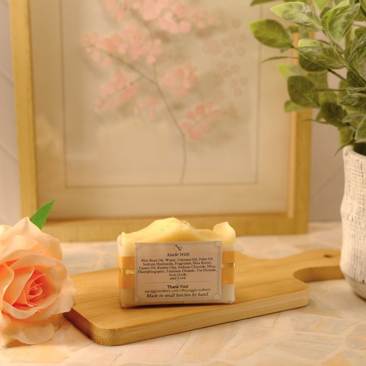 A bar of handmade soap sits on a wooden cutting board on a tiled countertop, styled with a peach rose, a potted green plant, and a framed picture in the background.