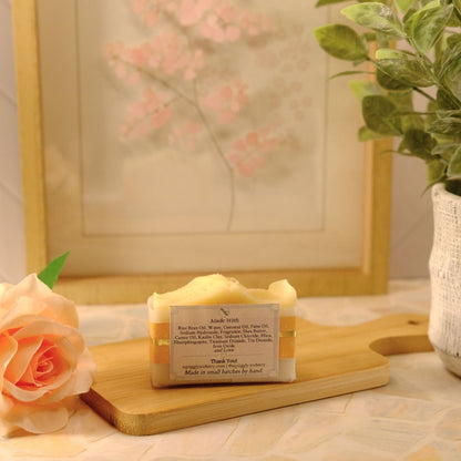 A bar of handmade soap sits on a wooden cutting board on a tiled countertop, styled with a peach rose, a potted green plant, and a framed picture in the background.